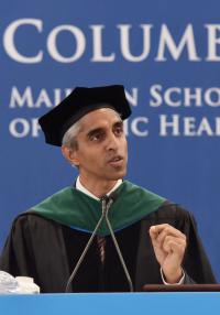 A man in academic robes speaks at a podium in front of a Columbia Mailman banner